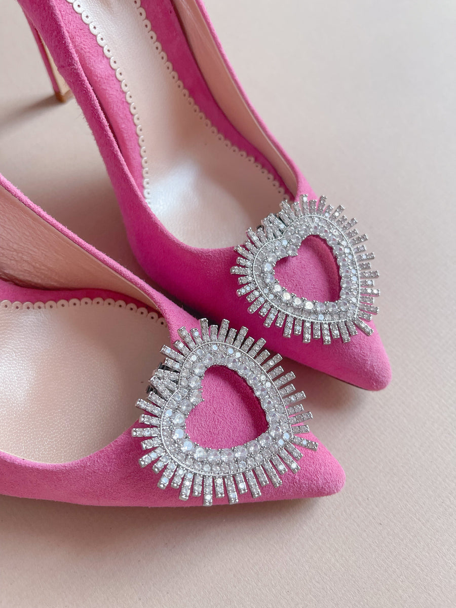 Pink suede heels featuring a prominent heart-shaped embellishment adorned with sparkling rhinestones. The image captures a close-up view of the shoes, highlighting the intricate detailing of the embellishment against a soft, neutral background. The angle showcases the pointed toe design and the elegant silhouette of the heels. image