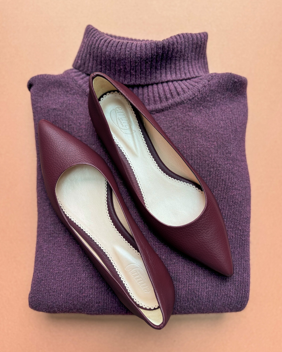 Elegant burgundy pointed-toe flats made of leather, displayed on a soft purple sweater. The image is taken from a top-down angle, showcasing the shoes' sleek design and smooth interior. The background is a warm, neutral color that complements the overall aesthetic. image