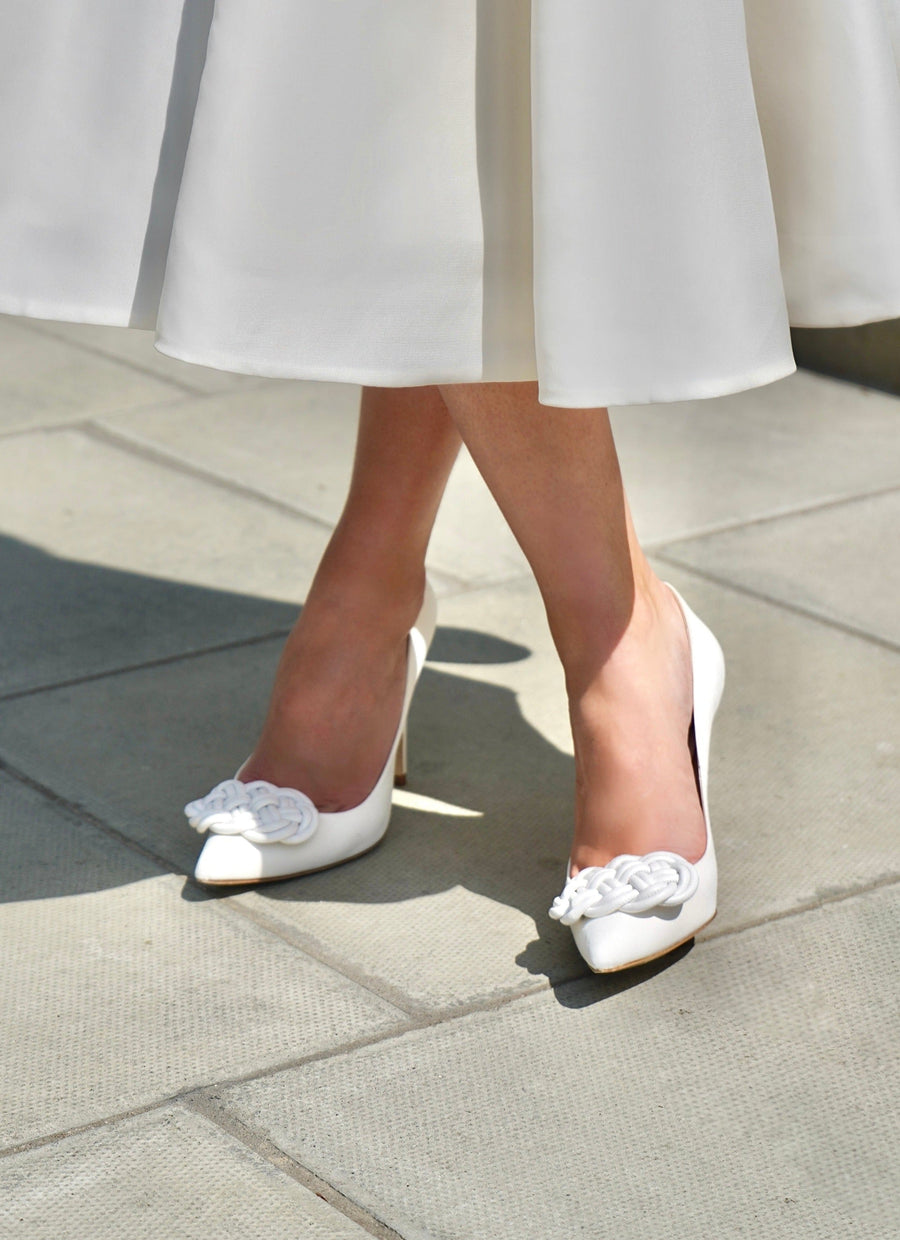 White pointed-toe heels with decorative floral embellishments at the front, worn by a person standing on a light-colored tiled surface. The heels are approximately 4 inches high, complementing a flowing white skirt that falls just below the knee. The image is taken from a low angle, highlighting the shoes against a bright outdoor background. image
