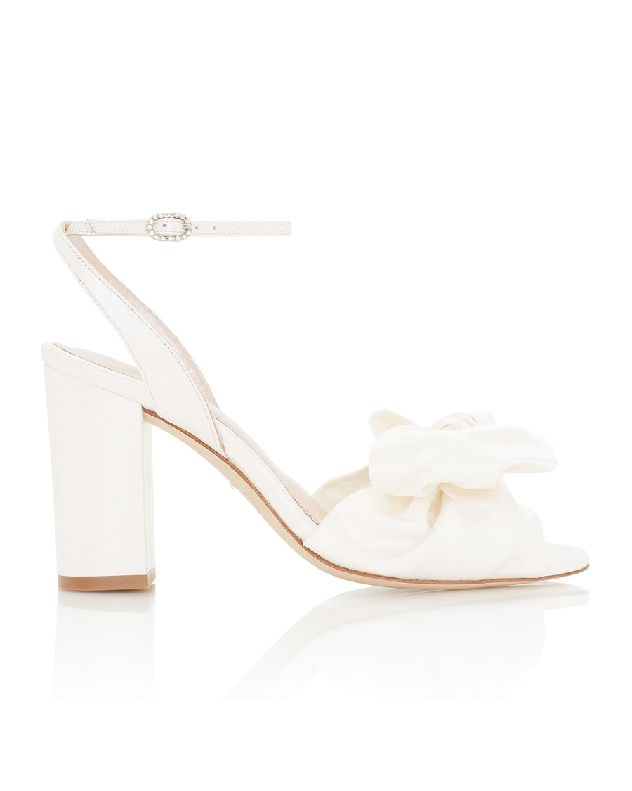 White heeled sandals featuring a wide strap adorned with a large bow on the front. The heels are block-style, providing stability, and the shoes have an ankle strap with a decorative buckle. The image is taken from a side angle against a clean, white background, showcasing the elegant design. image