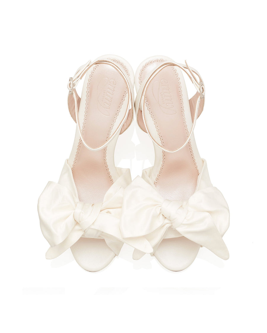 White sandals with large bow detail on the front, featuring a delicate ankle strap. The shoes are set against a clean white background, showcasing their elegant design. The heel height is approximately 3 inches. The material is likely leather. image