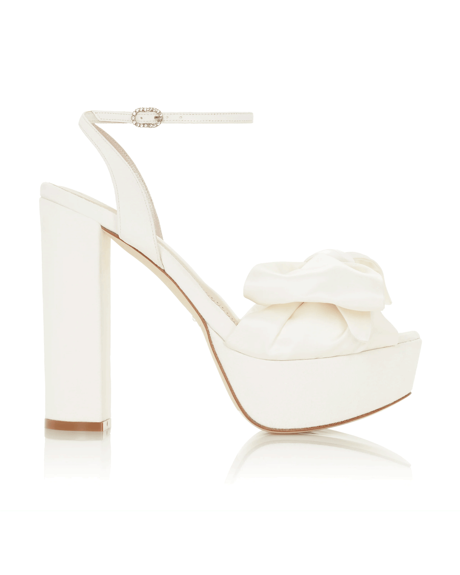 White platform heels with a large satin bow on the front. The shoes feature a chunky heel and an ankle strap for added support. The image is taken against a clean, white background, showcasing the shoes from a side angle. image