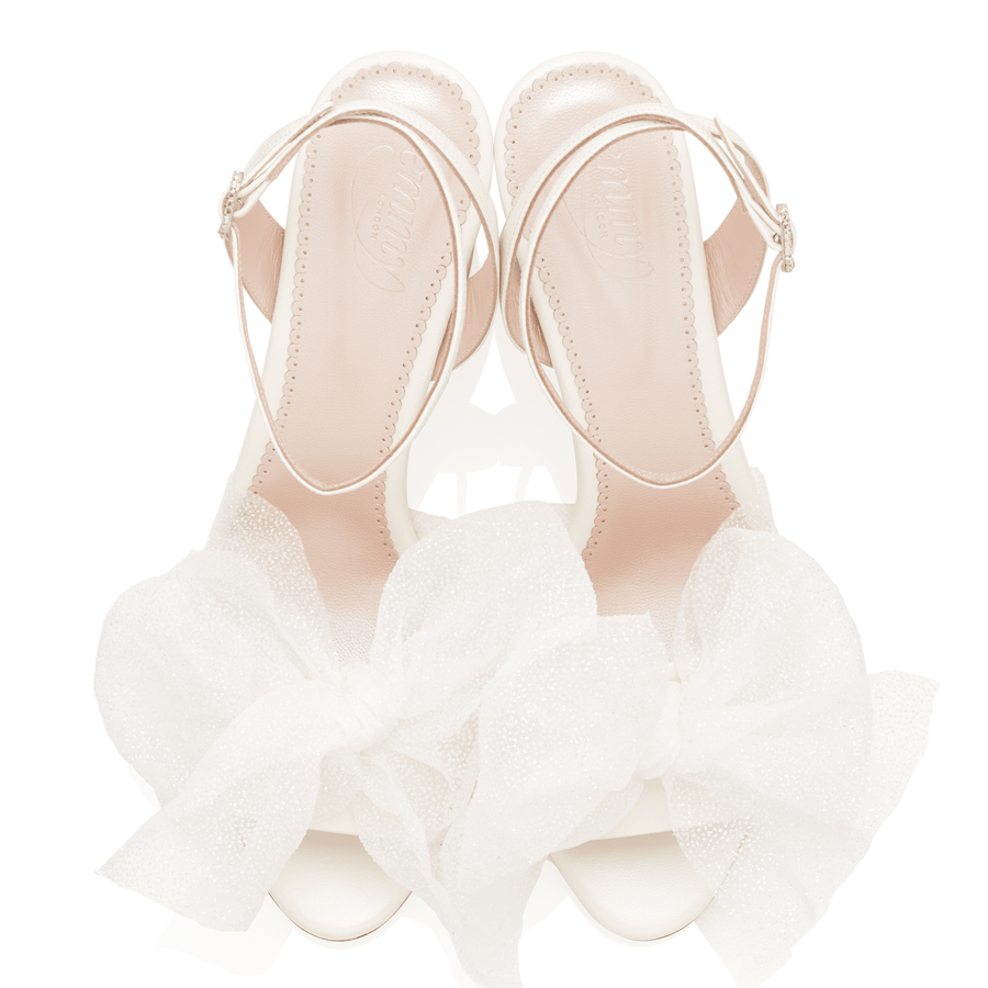 White heels with a delicate bow detail at the front, featuring a sleek design and an ankle strap. The shoes are positioned on a plain white background, showcasing their elegant silhouette. The angle captures the shoes from above, emphasizing the soft texture of the material and the intricate bow. image