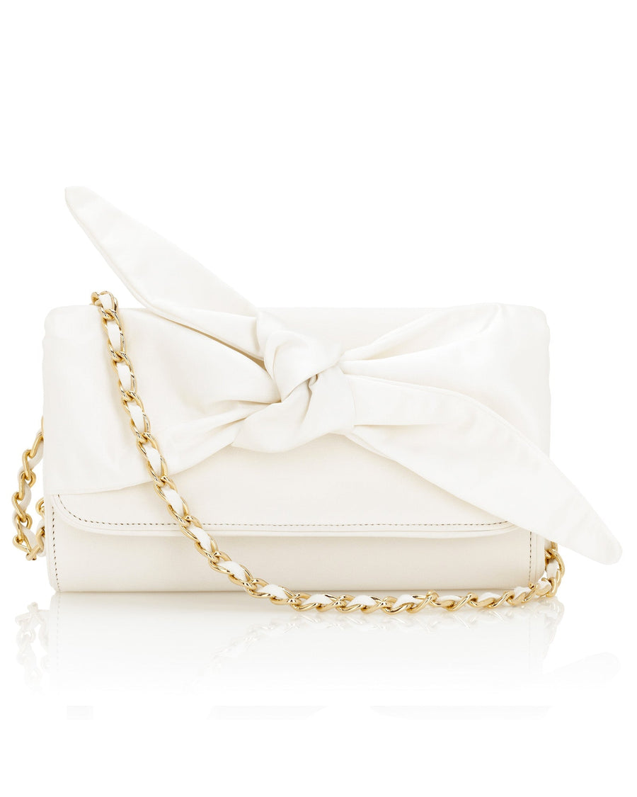 White handbag with a large bow detail on the front, featuring a gold chain strap. The bag is placed on a reflective surface, showcasing its smooth texture and elegant design. The background is neutral, allowing the bag to stand out prominently. image