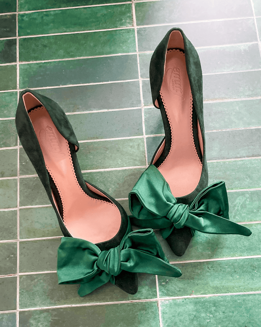 Green suede heels with a large bow detail at the front, set against a green tiled background. The shoes are positioned side by side, showcasing their pointed toes and elegant silhouette. image