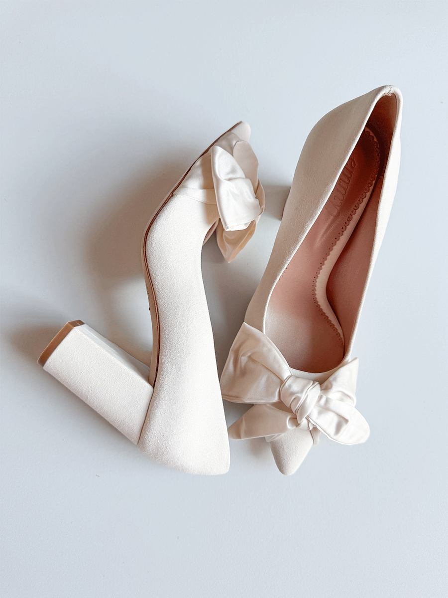 White block-heeled shoes with a decorative bow on the side, set against a light background. The shoes are positioned at an angle, showcasing the bow detail and the smooth texture of the material. image
