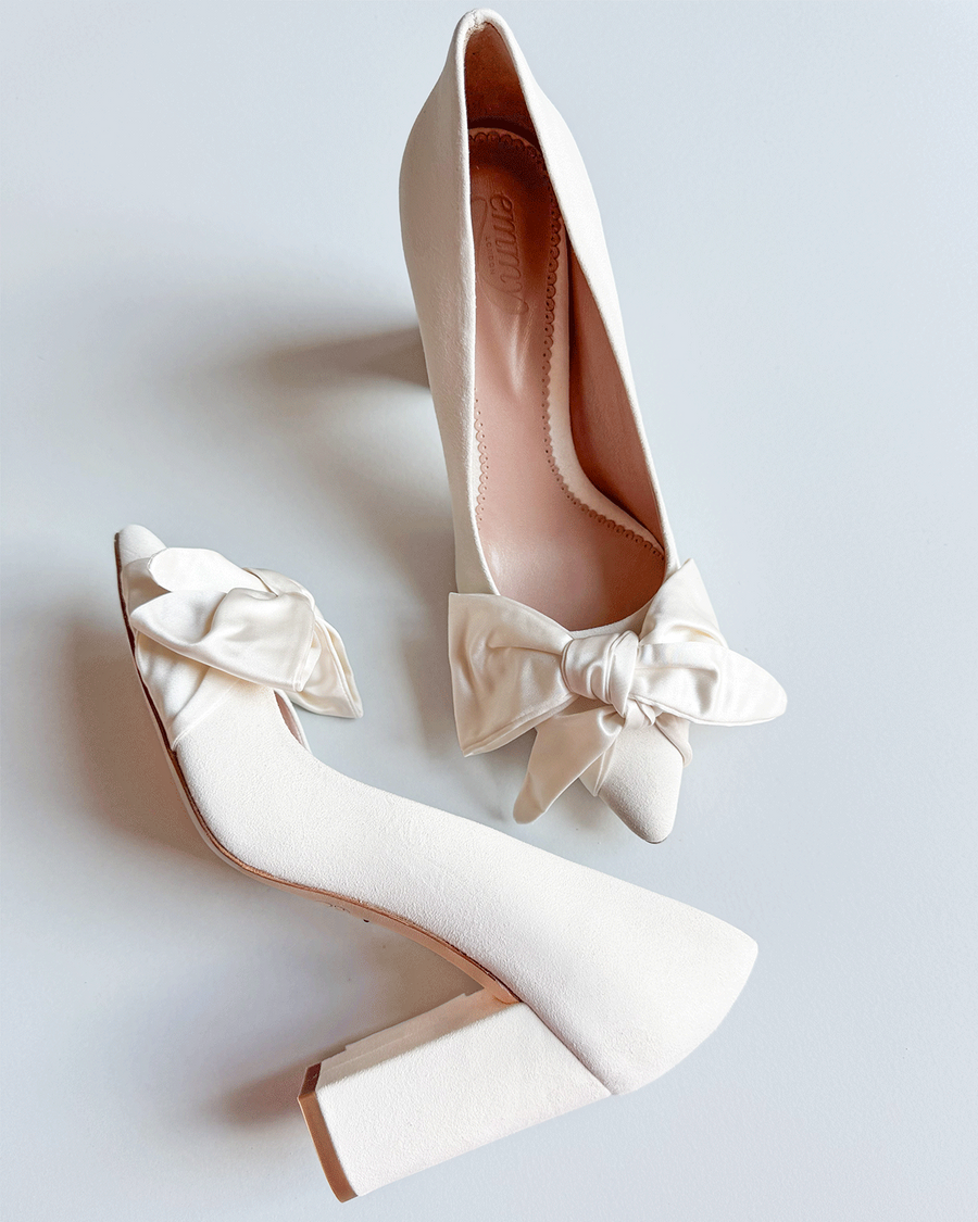 White heels with a chunky block heel and a large satin bow on the front. The shoes are positioned on a light gray background, showcasing their elegant design. The angle captures both the side and top view of the shoes, highlighting the smooth texture and clean lines. image
