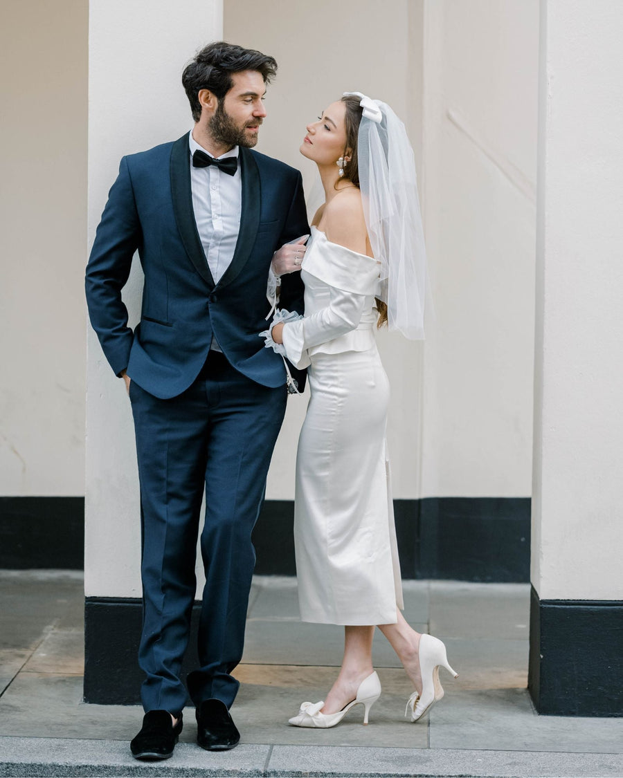 Elegant white heels with a moderate heel height complement a bride in a satin off-the-shoulder wedding dress, standing beside a groom in a tailored black tuxedo. The couple is positioned against a light-colored wall, creating a soft, romantic atmosphere. The bride's veil adds a delicate touch, while the groom's confident stance enhances the overall elegance of the scene. image