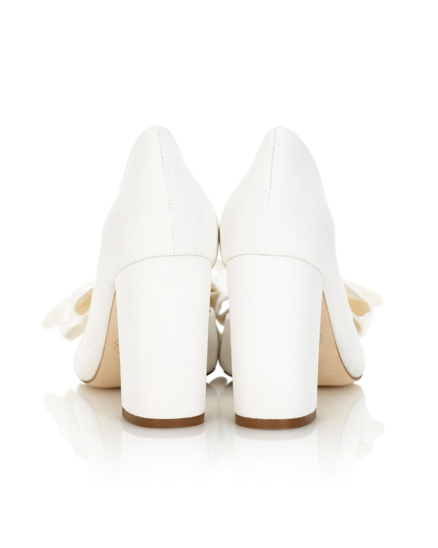 White block-heeled shoes with a smooth finish, featuring a bow detail at the back. The image is taken from a rear angle, showcasing the heels and the clean, reflective surface beneath them. The background is plain white, emphasizing the shoes. image
