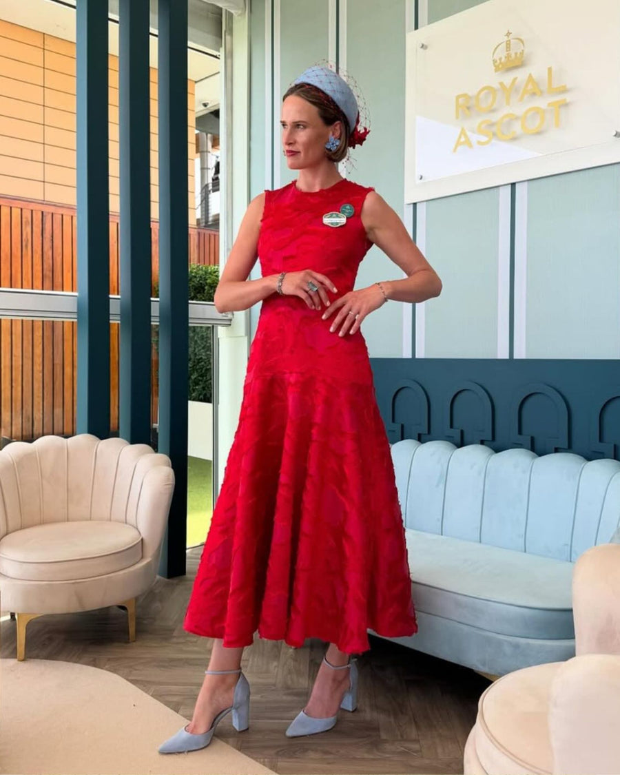 A woman stands elegantly in a bright red dress with a flared skirt, showcasing a pair of light blue heels. The image is taken indoors, with a stylish backdrop featuring a cream-colored sofa and a light blue chair. The setting is sophisticated, likely at a formal event. The heels are made of suede and have a moderate height, complementing her outfit perfectly. image