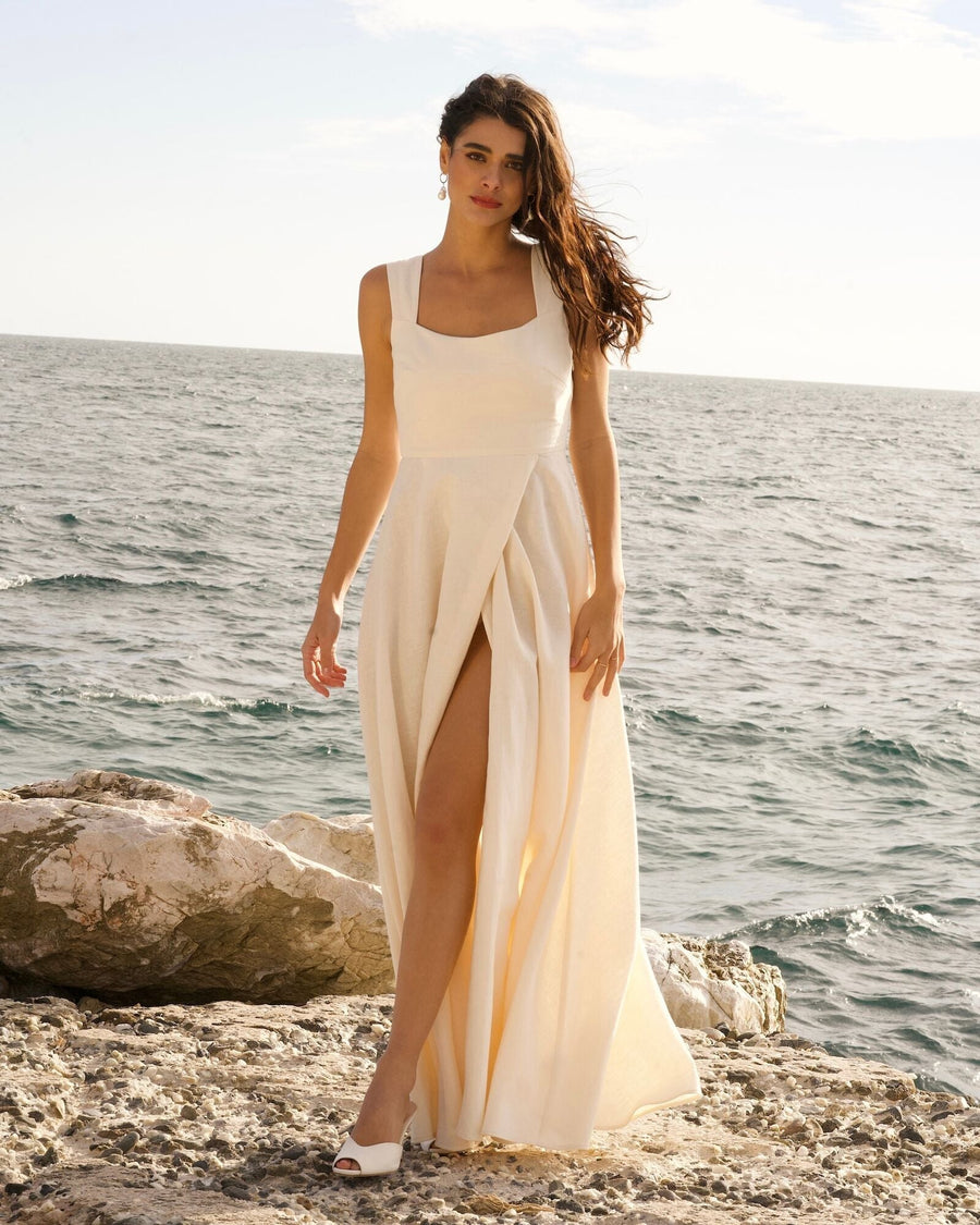 A woman stands on a rocky shoreline by the sea, wearing a flowing white dress with a thigh-high slit. She is barefoot, and the background features a calm ocean under a clear sky. The image captures her in a natural pose, with the wind gently tousling her hair. image