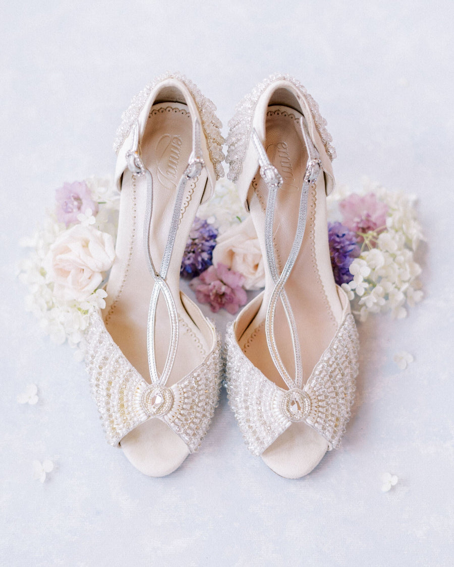 Elegant open-toe heels adorned with intricate beading and embellishments, set against a soft pastel background with delicate flowers surrounding them. The shoes feature a slender ankle strap and a stylish design, showcasing their luxurious appeal. image