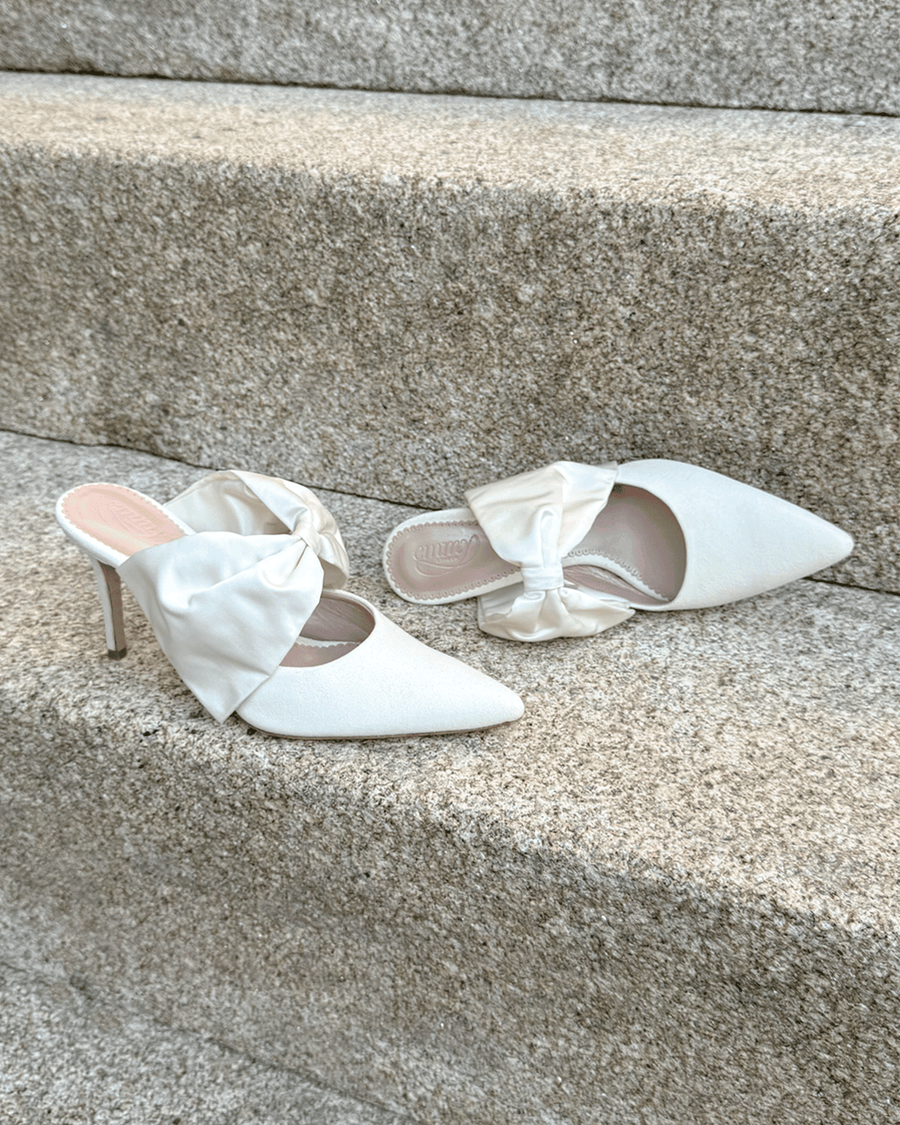 White pointed-toe heels with a decorative bow on the front, set against a textured stone background. The shoes are positioned on a staircase, showcasing their elegant design. The heels are approximately 3 inches high. image