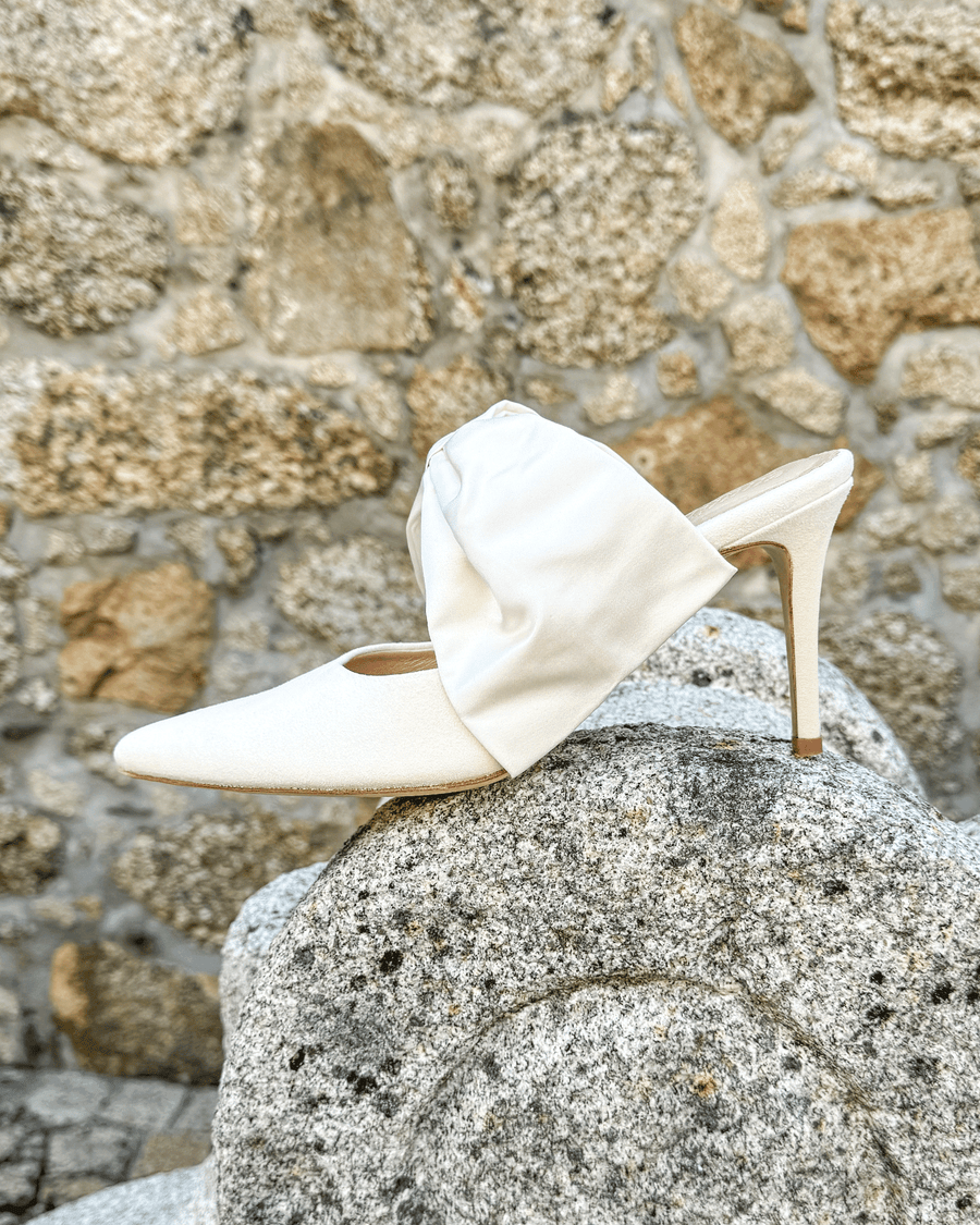 White pointed-toe heels with a fabric bow detail on the side, set against a textured stone background. The shoe features a stiletto heel, showcasing a sleek and elegant design. The image captures the shoe from a side angle, highlighting its shape and the contrast between the smooth fabric and the rough stone surface. image