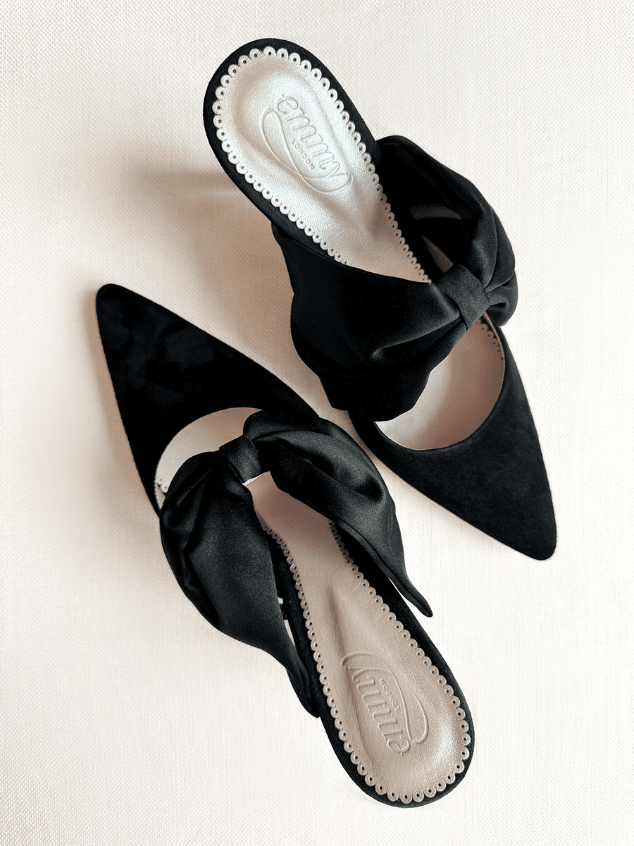 Black suede mules featuring a pointed toe and a large bow detail on the front. The shoes are positioned on a light textured background, showcasing their elegant design. The angle captures the top view, highlighting the smooth interior lining and the stylish silhouette. image