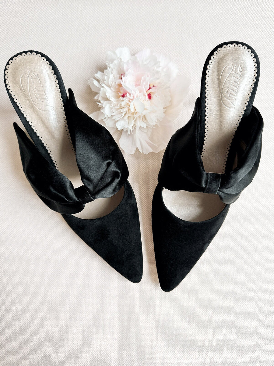 Black suede heels featuring a pointed toe and a decorative bow on the front. The shoes are positioned on a light-colored surface, with a soft pink flower placed nearby, creating a delicate and elegant aesthetic. The image is taken from a top-down angle, showcasing the intricate details of the shoes. image