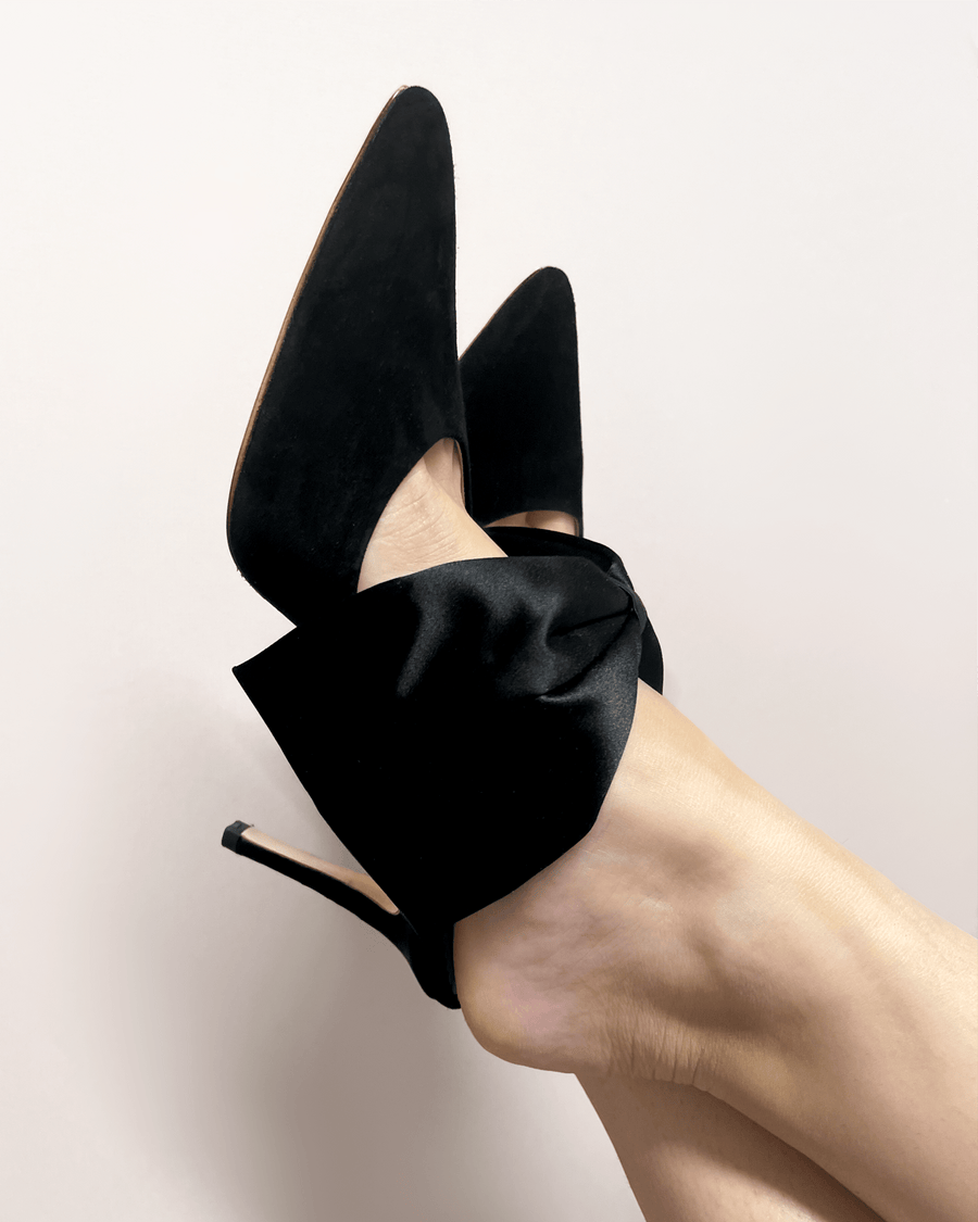 Black suede heels with a pointed toe and a large black satin bow around the ankle. The shoes are being worn by a person whose foot is positioned at an angle, showcasing the heel height, which is approximately 4 inches. The background is a soft, neutral color, enhancing the elegance of the footwear. image