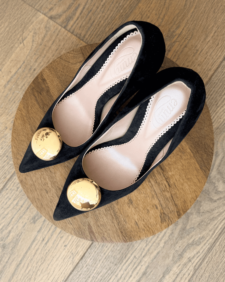Black suede pointed-toe heels with a gold embellishment on the vamp, displayed on a wooden round surface. The image is taken from a top-down angle, showcasing the shoes without any model. The background features a light wooden floor. image