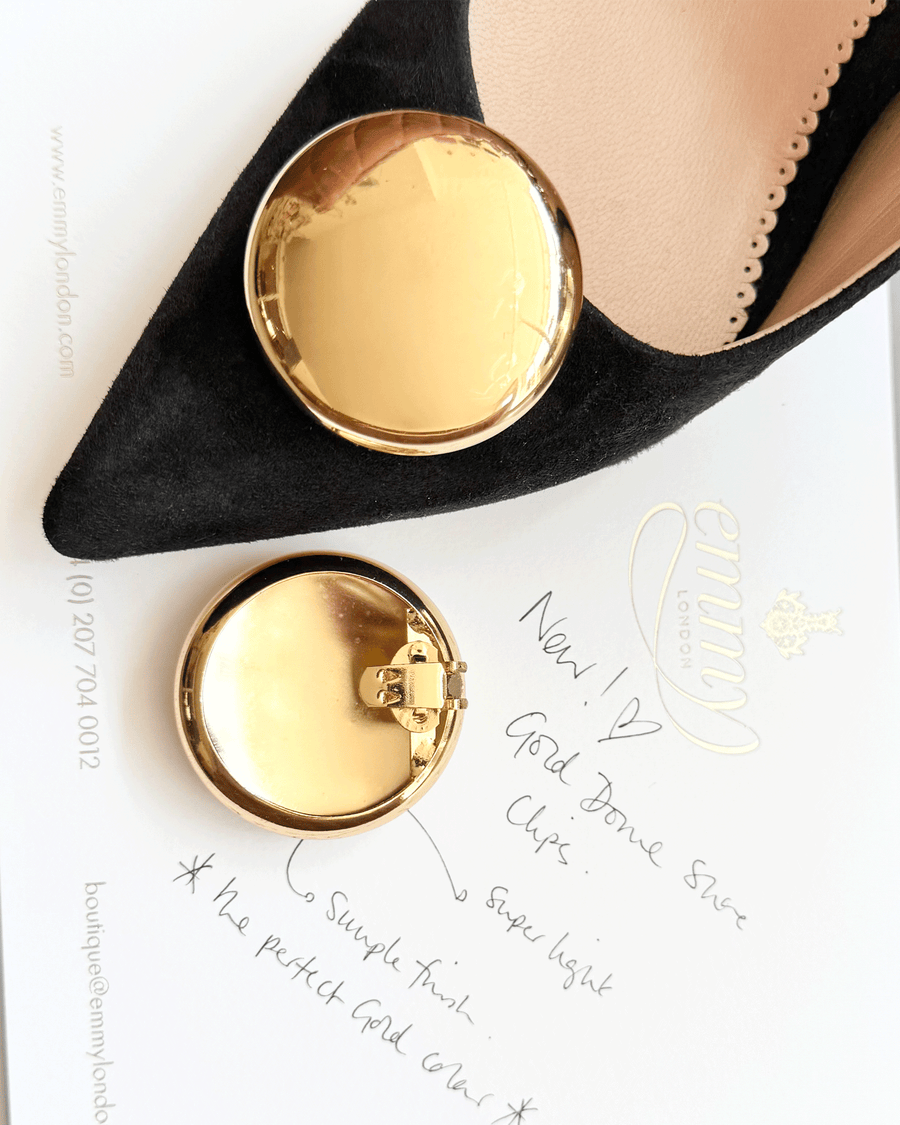 Black suede heels featuring a pointed toe and a large gold circular embellishment on the front. The image is taken from a top-down angle, showcasing the shoe alongside a gold-tone round clasp. The background includes a white card with handwritten notes, emphasizing the elegant presentation of the product. image