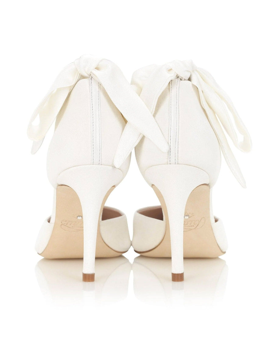 White heels with a sleek design featuring a satin bow at the back. The shoes have a pointed toe and a stiletto heel, positioned on a reflective surface that enhances their elegance. The background is neutral, allowing the shoes to stand out prominently. image