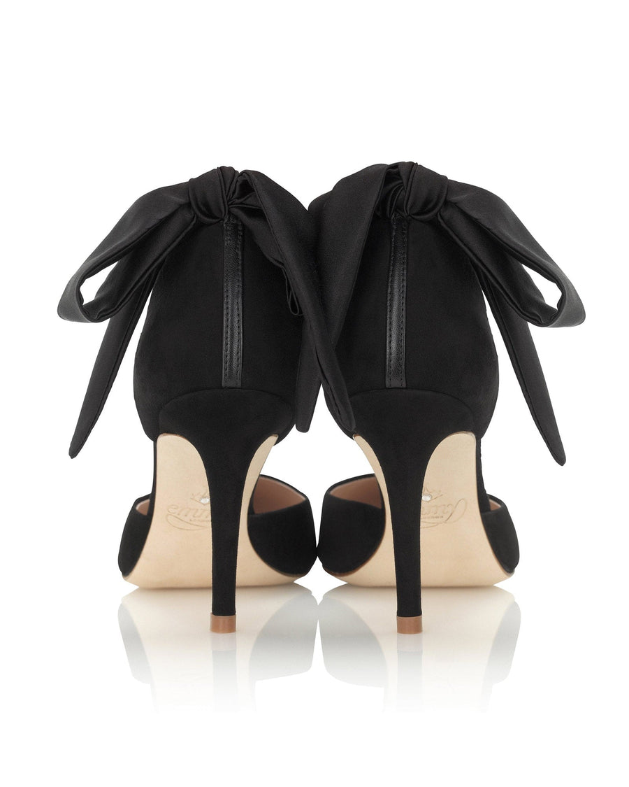 Black suede heels featuring a large satin bow at the back. The shoes have a sleek design with a pointed toe and a stiletto heel. The image is taken against a clean, white background, showcasing the shoes from a rear angle. image