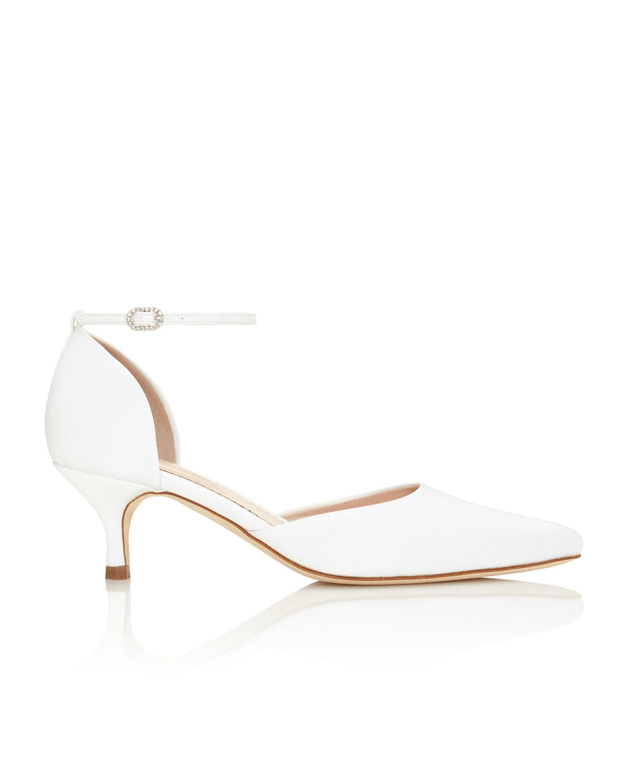 White pointed-toe heels with a delicate ankle strap, featuring a modest heel height. The shoes are set against a clean, bright background, showcasing their elegant design. The angle highlights the sleek silhouette and refined details of the footwear. image
