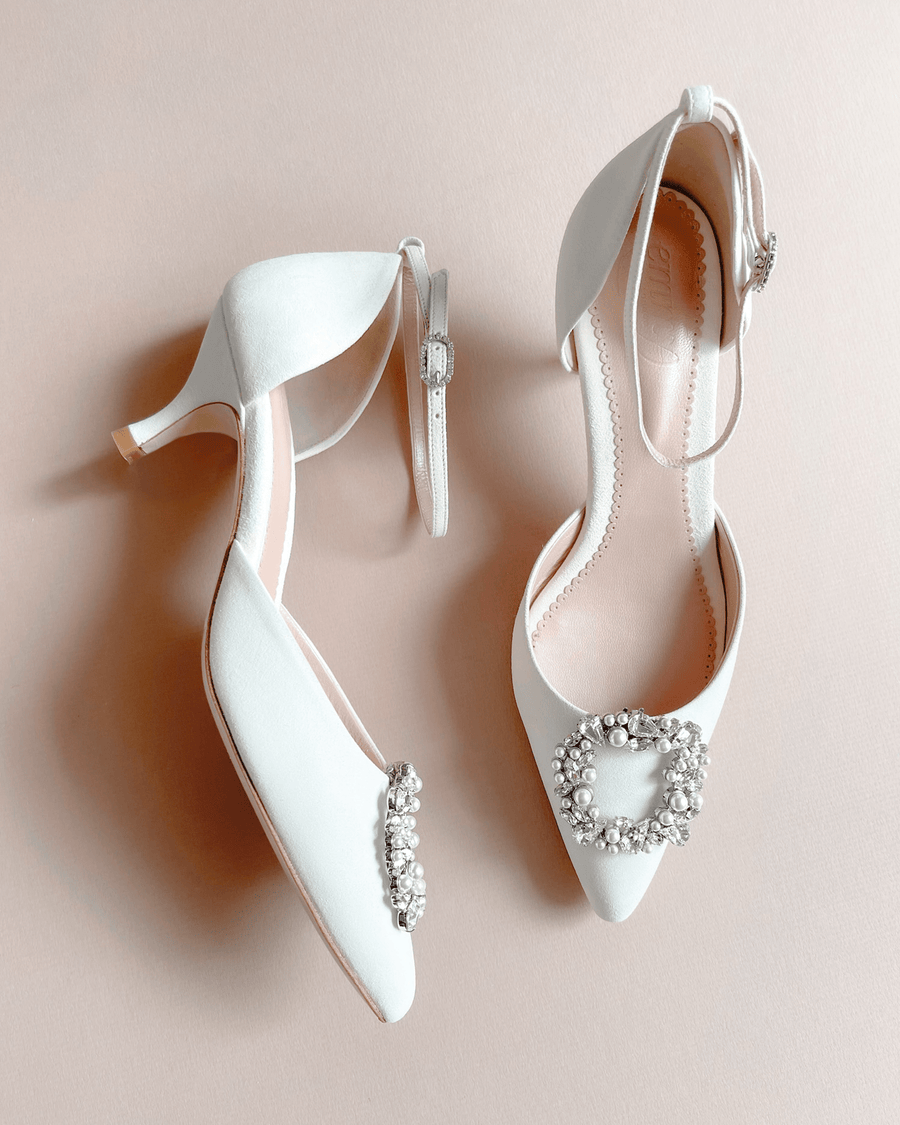 White leather heels with a pointed toe and a decorative crystal buckle on the front. The shoes are displayed on a soft beige background, showcasing their elegant design. The angle captures both shoes side by side, highlighting the delicate ankle strap on one shoe. image