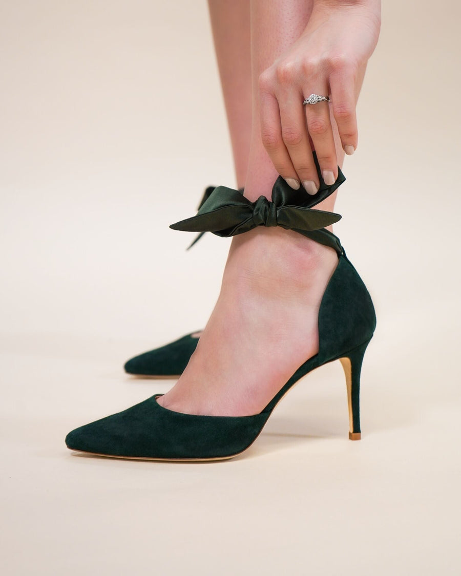 Green suede heels with a pointed toe and a slender stiletto heel. The model is adjusting a satin ribbon tied around the ankle, set against a soft beige background. The angle showcases the shoe's elegant silhouette and the delicate detailing of the ribbon. image
