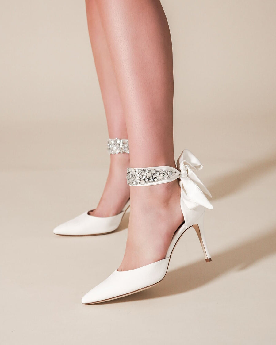 White pointed-toe heels with a delicate ankle strap adorned with sparkling embellishments, worn by a model standing against a soft beige background. The heels are approximately 3 inches high. image