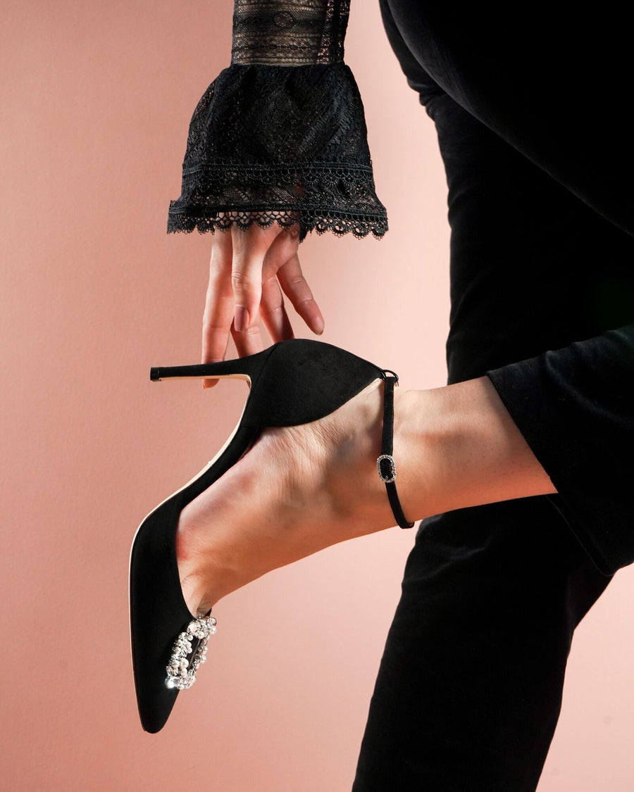 Black suede heels with a pointed toe and a decorative brooch on the front. The shoe is being held by a person wearing a black lace sleeve, with a soft pink background. The heel height is approximately 4 inches. image