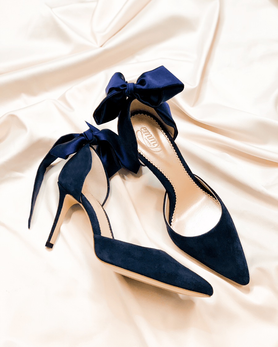 Navy blue suede heels with a pointed toe and a delicate bow detail at the ankle. The shoes are positioned on a soft, cream-colored satin fabric, showcasing their elegant design. The image captures the shoes from a slightly elevated angle, highlighting the sleek silhouette and the luxurious texture of the material. image
