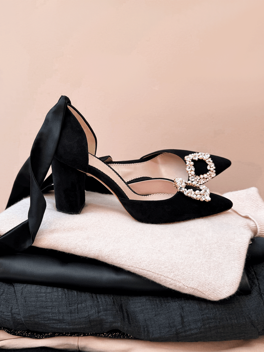Black suede heels with a chunky block heel, featuring a decorative buckle adorned with pearls. The shoes are positioned on a stack of folded clothing, with a soft pink background that complements the overall aesthetic. The image is taken from a top-down angle, showcasing the elegant design and detailing of the shoes. image