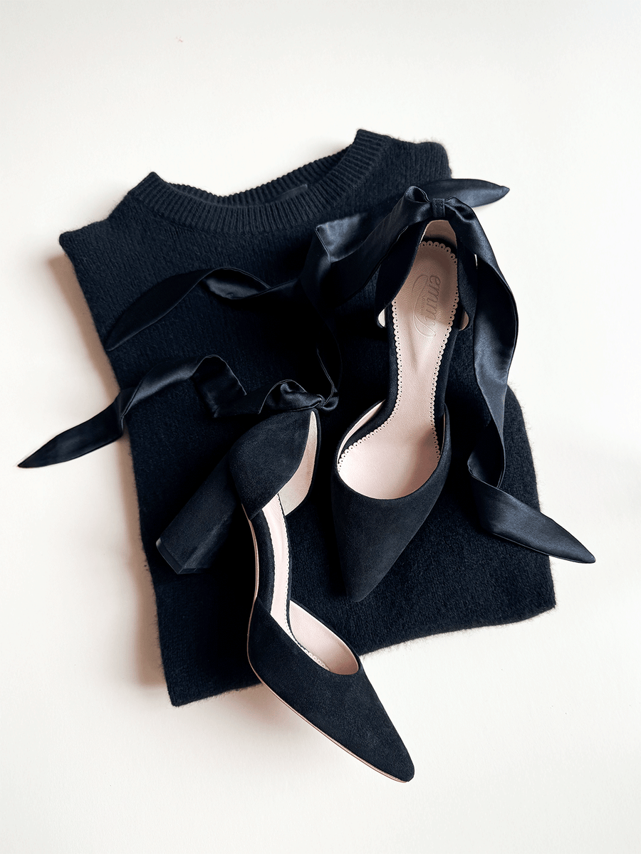 Black suede heels with a chunky heel are positioned on a soft black sweater. The shoes feature elegant satin ribbons tied around the ankle, adding a stylish touch. The image is taken from a top-down angle, showcasing the shoes against a neutral background. image