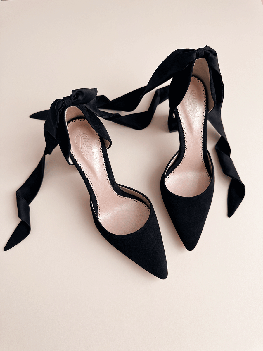 Black suede high heels with a pointed toe and elegant satin ribbons tied at the ankle. The shoes are positioned on a light beige surface, showcasing their sleek design. The image is taken from a top-down angle, emphasizing the delicate details of the ribbons and the smooth interior of the shoes. image