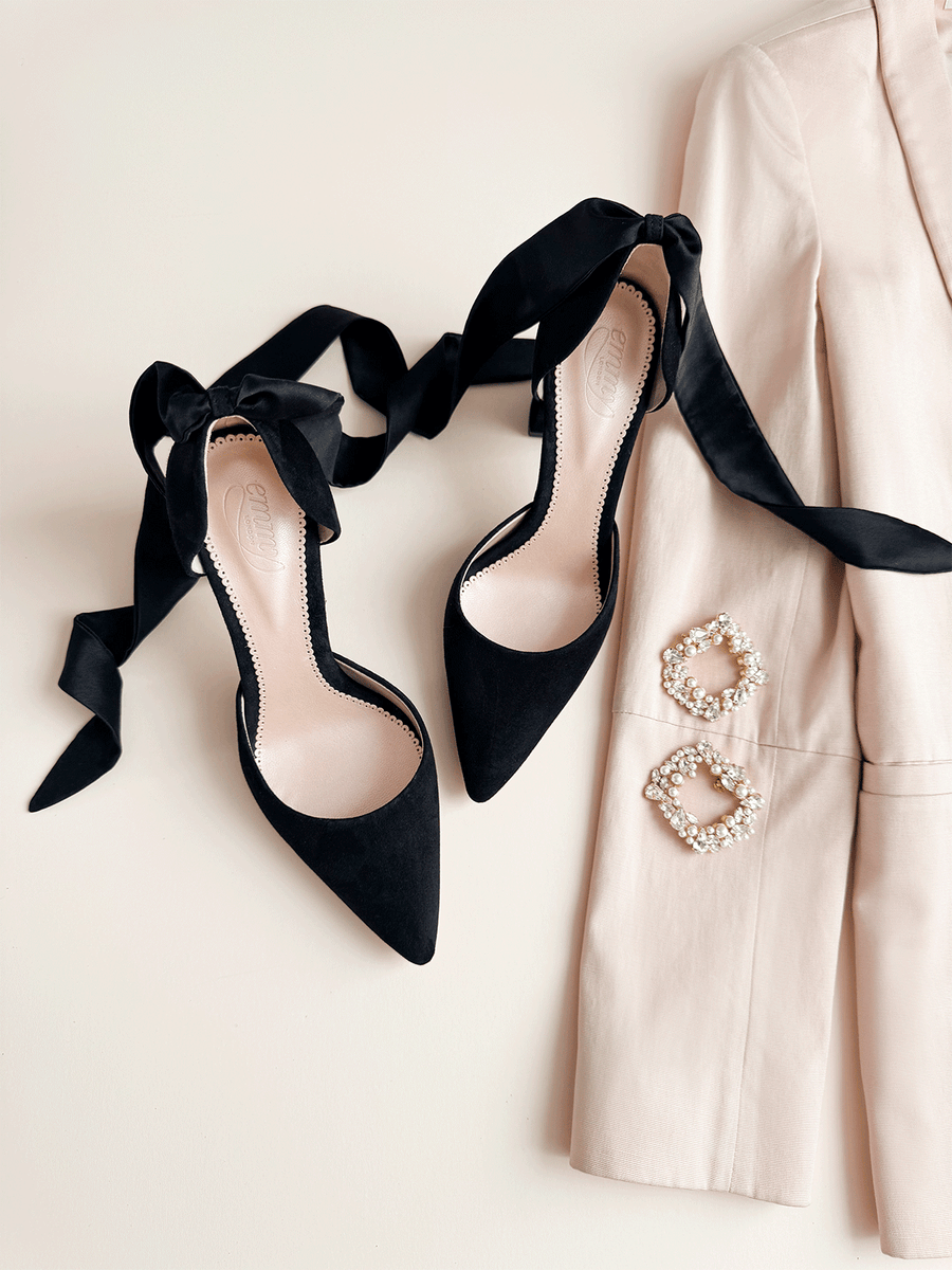 Black suede heels with a pointed toe design are displayed alongside a light-colored blazer and two decorative brooches. The shoes feature elegant black satin ribbons tied around the ankle, adding a sophisticated touch. The image is taken from a top-down angle, showcasing the shoes against a neutral background. image