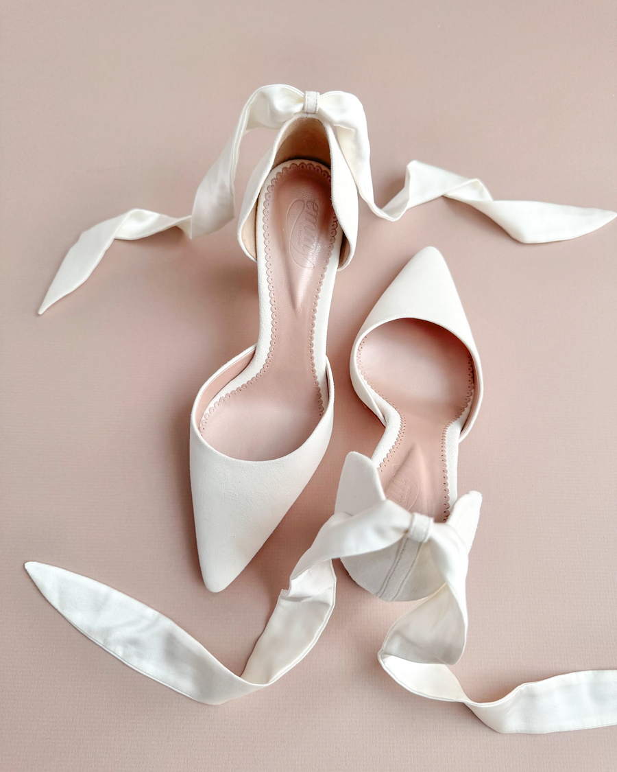 White pointed-toe heels with satin ribbon ties, displayed on a soft pink background. The shoes are positioned at an angle, showcasing their elegant design and smooth finish. image