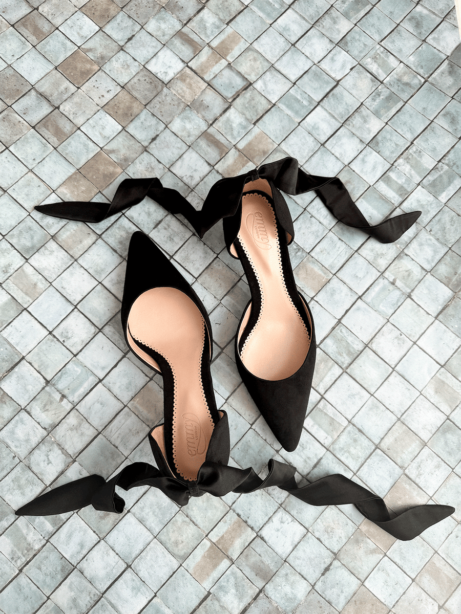 Black pointed-toe heels with a sleek design, featuring satin ribbon ties at the ankle. The shoes are displayed on a textured light blue and gray tiled surface, showcasing their elegant silhouette. The angle captures the shoes from above, emphasizing the sharp toe and the delicate ribbon detail. image
