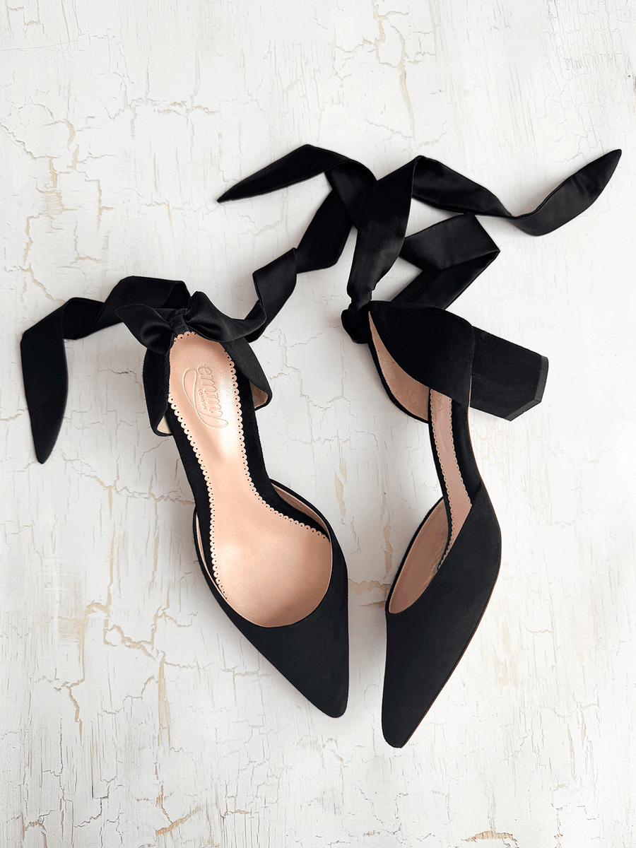Black suede heels with a block heel, featuring elegant satin ribbons tied around the ankle. The shoes are positioned on a textured white background, showcasing their pointed toe design and open sides. image