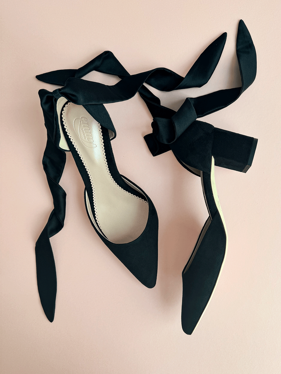Black suede heels with a square toe and a chunky heel, featuring elegant black satin ribbons tied around the ankle. The shoes are displayed against a soft pink background, showcasing their sleek design from a top-down angle. image