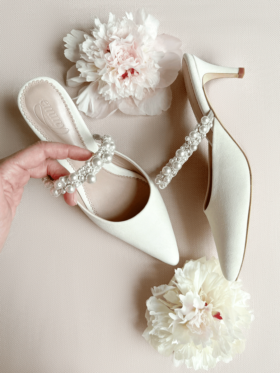 Elegant white heels with a pointed toe design, featuring a decorative pearl strap across the front. The shoes are made of a soft material and are positioned on a light pink background, accompanied by large, delicate peonies. A hand is gently holding one of the shoes, showcasing its stylish design and the intricate detailing of the pearl embellishments. The heel height is approximately 2.5 inches. image
