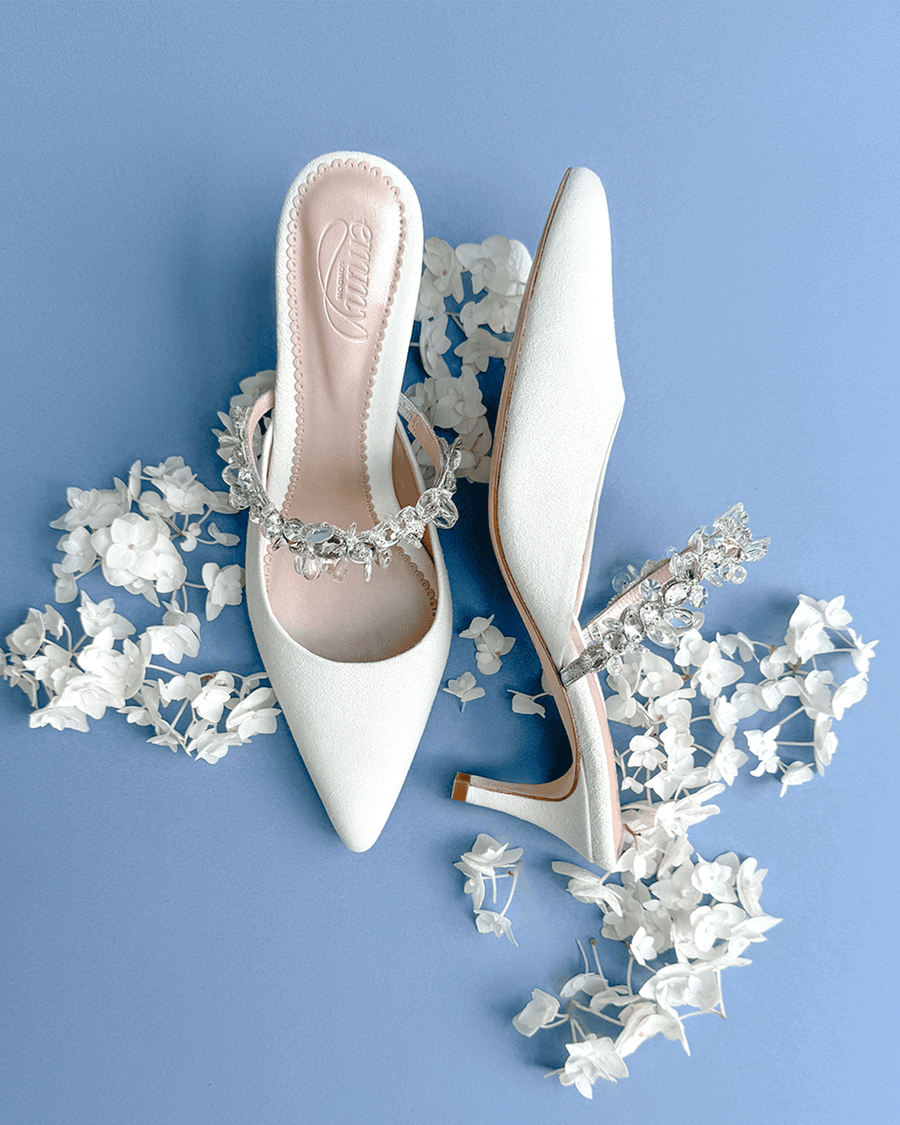 White pointed-toe heels with a decorative crystal strap, set against a light blue background. The shoes are positioned with one facing upward and the other slightly angled, surrounded by delicate white flower petals. The heel height is approximately 2.5 inches. image