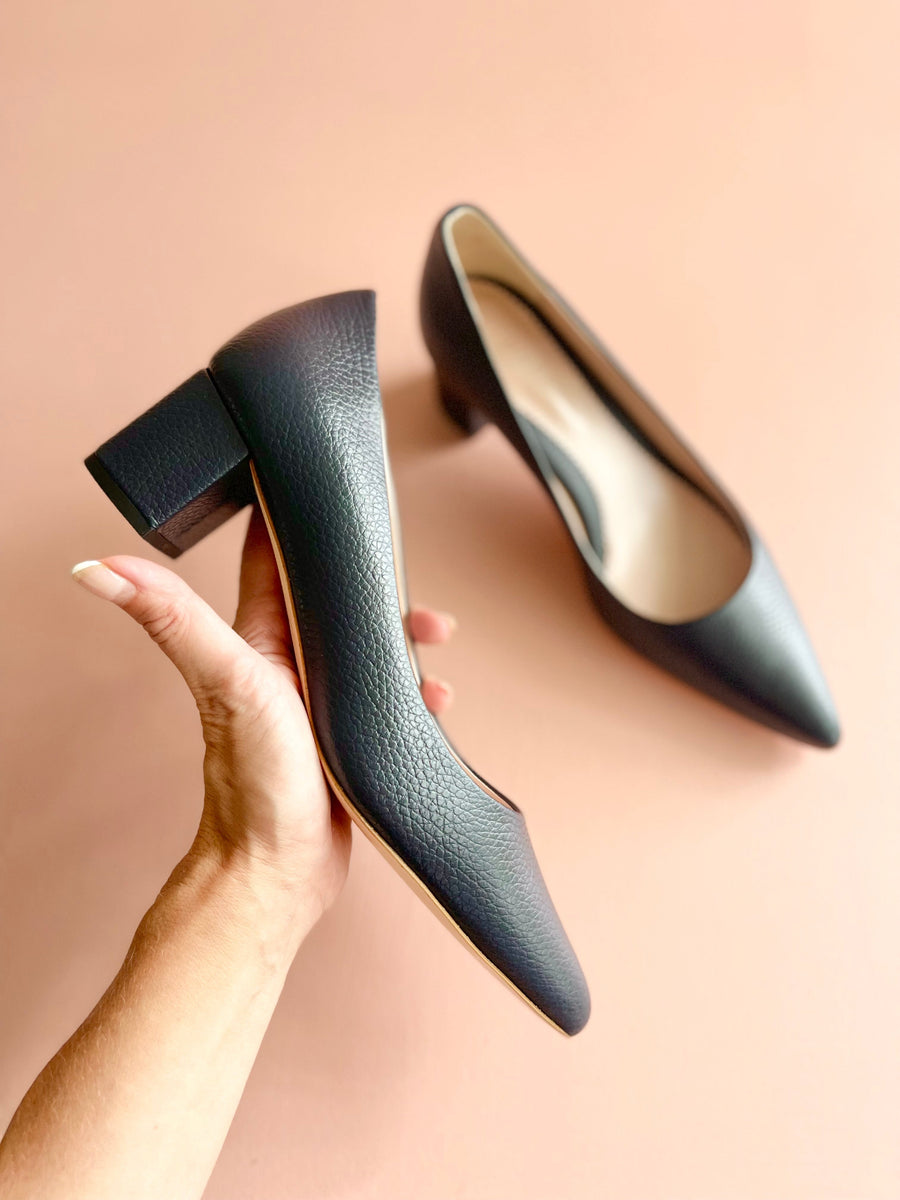 Navy leather heels with a mid-height block heel, held in a hand against a soft pink background. The shoes feature a pointed toe and a smooth texture, showcasing their elegant design. image