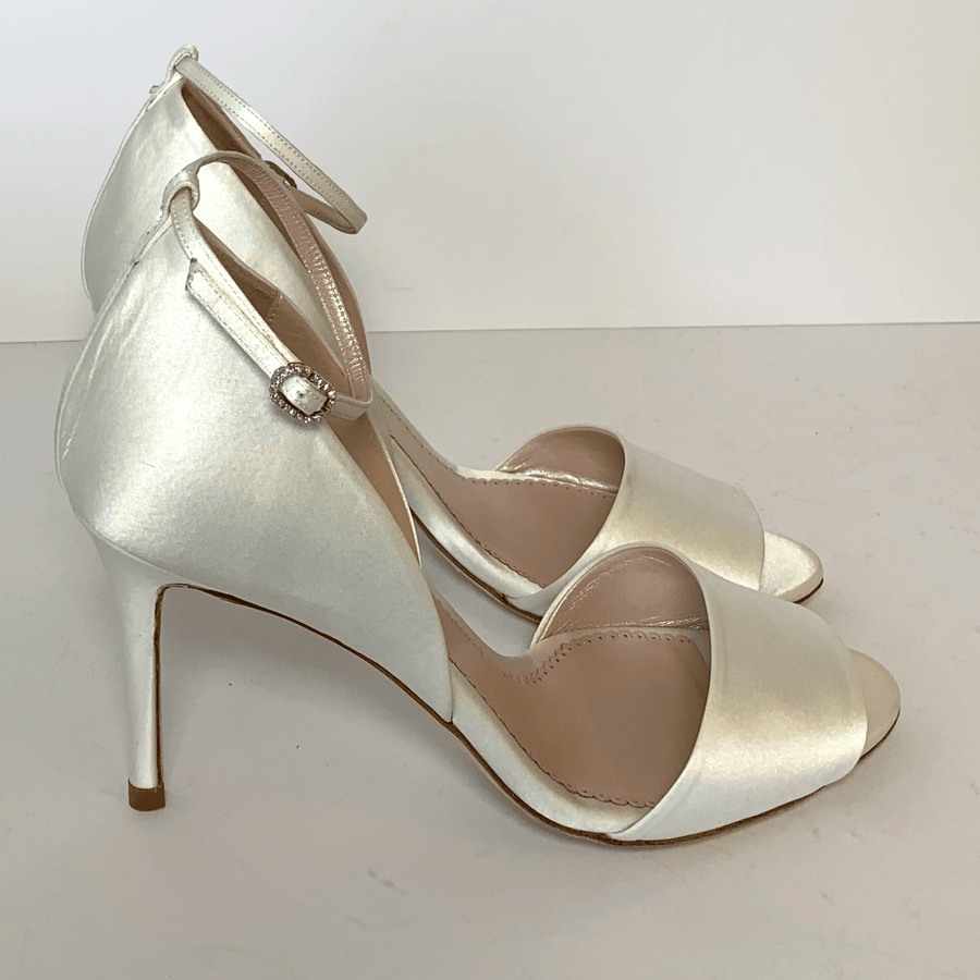 Elegant silver heels featuring a sleek design with a delicate ankle strap and a subtle embellishment. The shoes have a pointed toe and a stiletto heel, positioned on a clean, white background, showcasing their refined silhouette. image