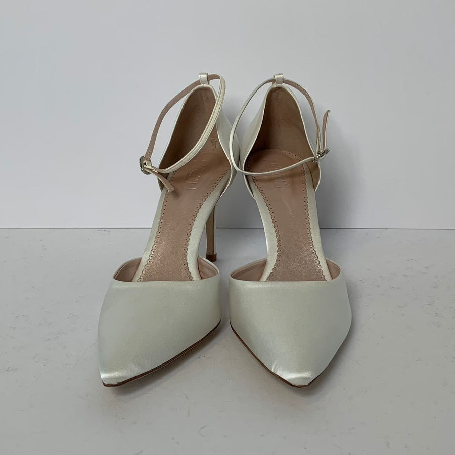 White pointed-toe heels with a slender ankle strap, set against a plain white background. The shoes feature a glossy finish and a stiletto heel, showcasing a modern and elegant design. image