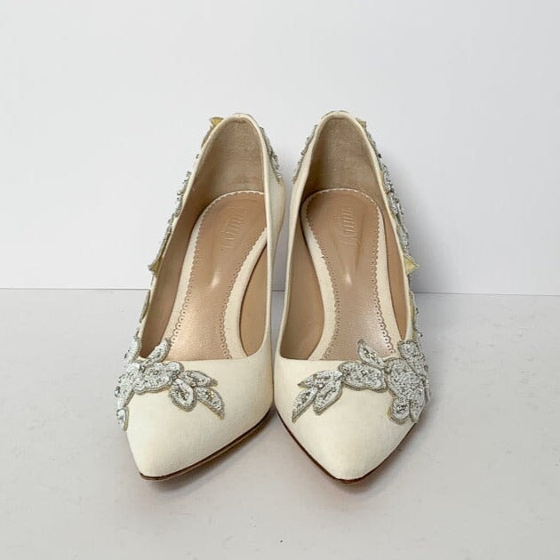 Elegant white heels adorned with intricate floral embellishments, set against a plain white background. The shoes feature a pointed toe design and a sleek silhouette, showcasing their refined style. image