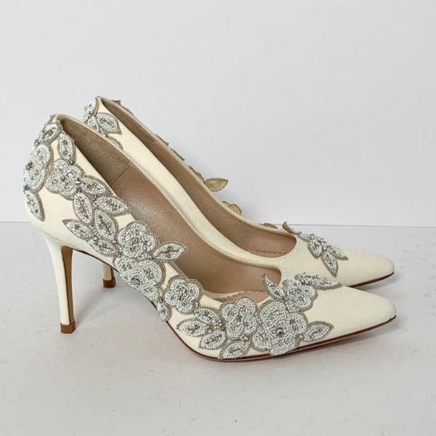 Elegant white heels adorned with intricate floral embellishments in silver. The shoes feature a pointed toe and a slender stiletto heel, set against a plain white background. The angle showcases the side view, highlighting the detailed design and craftsmanship. image