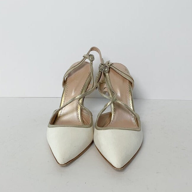 White pointed-toe heels with a delicate crisscross strap design, set against a plain white background. The shoes feature a mid-height heel and are made of suede. The image captures the shoes from a frontal angle, showcasing their elegant silhouette. image