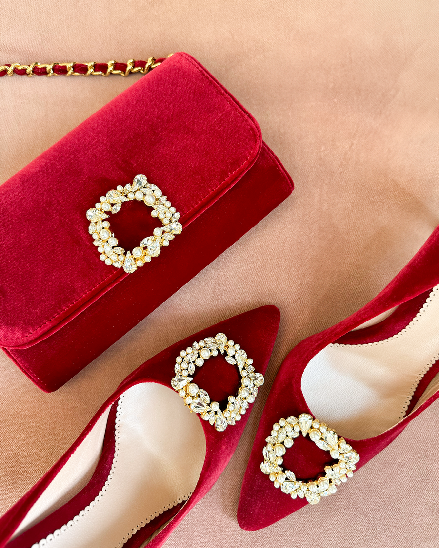 Red suede heels with a pointed toe and embellished buckle detail are positioned alongside a matching red velvet handbag. The image is taken from a top-down angle, showcasing the shoes and bag against a soft, neutral background. The heels are elegantly designed, perfect for a sophisticated look. image