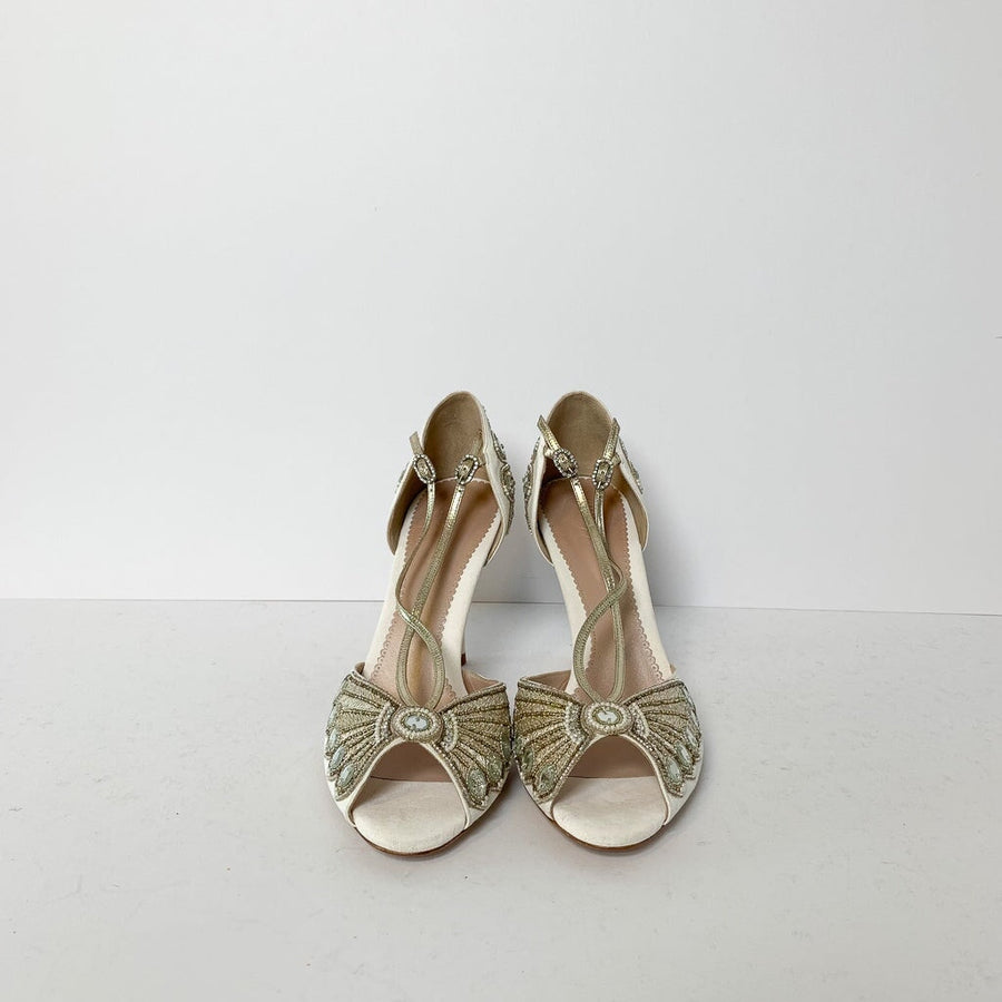 Elegant open-toe heels featuring intricate beading and embellishments in a soft cream color. The shoes have a delicate t-strap design and a slender heel. They are displayed against a clean, white background, showcasing their detailed craftsmanship. image