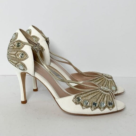 Elegant white heels featuring intricate gold and crystal embellishments on the front. The shoes have a delicate strap design and a slender stiletto heel. The image is taken from a side angle against a plain white background, showcasing the detailed craftsmanship of the shoes. image